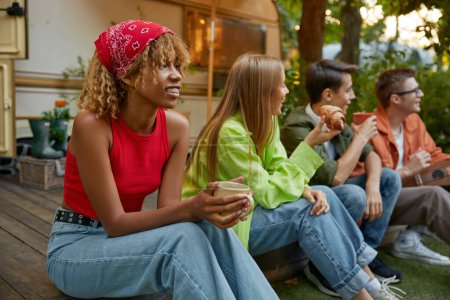 Photo for Happy multicultural group of young people, best friends team sitting in row eating snacks and drinking refreshment tea enjoying rest during picnic at campsite. Happiness and carefree summer time - Royalty Free Image