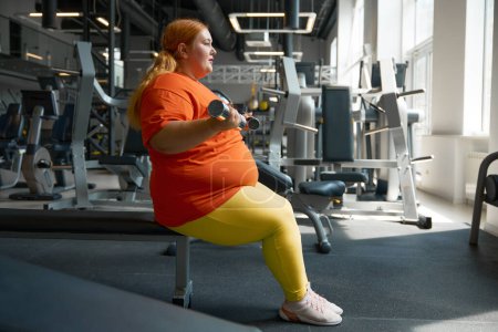 Téléchargez les photos : Femme de taille plus faire de l'haltérophilie exercice sur biceps d'entraînement avec haltères à la salle de gym. Concept positif du corps - en image libre de droit