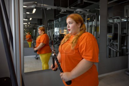 Photo for Exhausted excess overweight woman training with equipment at gym. Lovely pretty obese girl doing physical exercise for arms, shoulders and back. Sorts everyday routine for weightloss - Royalty Free Image