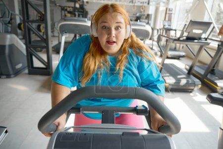 Téléchargez les photos : Portrait de femme en surpoids motivée faisant du vélo d'appartement au gymnase. routine sportive quotidienne pour la santé et la perte de poids - en image libre de droit