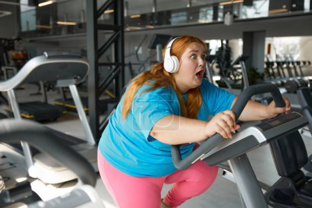 Téléchargez les photos : Portrait de femme en surpoids motivée faisant du vélo d'appartement au gymnase. routine sportive quotidienne pour la santé et la perte de poids - en image libre de droit