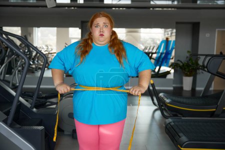 Foto de Retrato linda mujer divertida con exceso de peso ata a sí misma con cinta métrica de pie en el gimnasio. Dieta para adelgazar y entrenamiento deportivo para mujeres obesas - Imagen libre de derechos