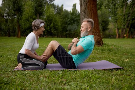 Photo for Happy married couple of retirees doing training workout in park. Elderly wife helping senior husband doing abdominal crunch. Sports leisure activity for pensioner - Royalty Free Image