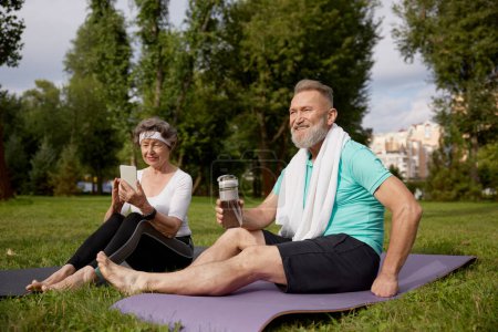 Téléchargez les photos : Couple âgé assis sur des tapis de fitness se reposer après l'entraînement de yoga en plein air dans le parc. Femme âgée utilisant un smartphone, homme mature buvant de l'eau. Week-end sportif et temps libre à la retraite - en image libre de droit