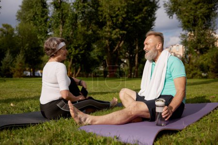 Photo for Senior couple sitting on fitness mats rest after yoga training outdoors in city park. Elderly woman using smartphone, mature man drinking water. Sports weekend and free time on retirement - Royalty Free Image