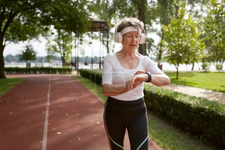 Téléchargez les photos : Femme âgée courant dans le parc tôt le matin. Sportive âgée portant un casque regardant bracelet suivi de remise en forme soigner sa santé - en image libre de droit