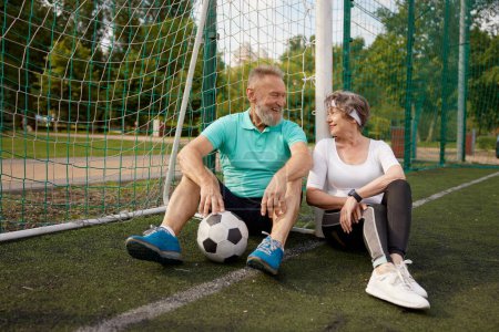 Photo for Happy smiling and tired seniors couple resting after successful soccer training. Married elderly man and woman sitting on field grass looking at each other with love and gratitude - Royalty Free Image