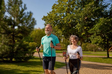 Téléchargez les photos : Joyeux couple retraité profitant d'une formation de marche en pole dans le parc municipal. Homme et femme âgés portant des vêtements de sport faisant de l'exercice à l'extérieur - en image libre de droit