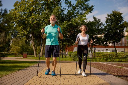 Foto de Feliz pareja jubilada disfrutando del entrenamiento de pole walk en el parque público urbano. Anciano y mujer usando ropa deportiva haciendo ejercicio al aire libre - Imagen libre de derechos