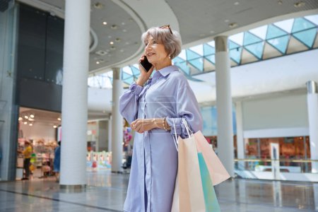 Photo for Portrait of attractive senior woman with phone and shopping bags over mall store showcase. Stylish grandmother feeling satisfied enjoying discounts in shop - Royalty Free Image