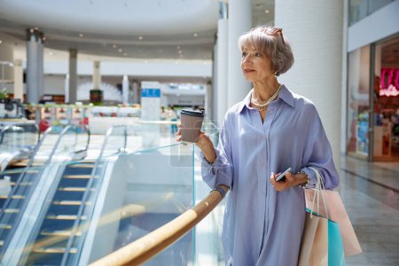 Téléchargez les photos : Femme âgée avec tasse de café et sacs à provisions sur vitrine du centre commercial. Élégant grand-mère se sentant satisfait profiter de réductions dans la boutique - en image libre de droit