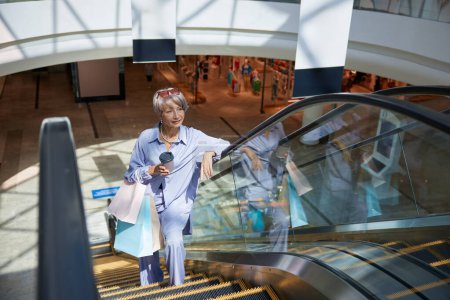 Téléchargez les photos : Heureuse femme retraitée chevauchant l'escalator au centre commercial anticipant de grandes réductions et des ventes sur Black Friday - en image libre de droit