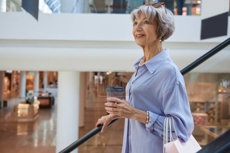 Photo for Aged woman in casual clothes with takeaway coffee on escalator. Shopping time on pension concept - Royalty Free Image