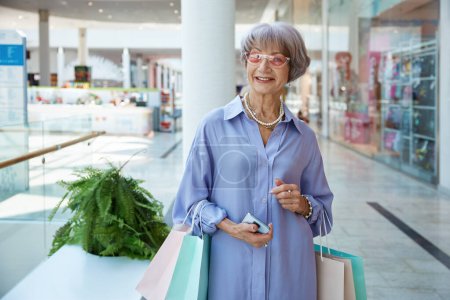 Foto de Mujer mayor atractiva feliz con bolsas de compras sobre escaparate de la tienda del centro comercial. Elegante abuela sintiéndose satisfecho disfrutando de descuentos en tienda - Imagen libre de derechos