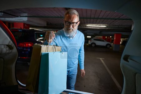 Foto de Senior comprador masculino cargando coloridas bolsas de comprador en el maletero del coche. Consumismo y compras diarias concepto de rutina - Imagen libre de derechos