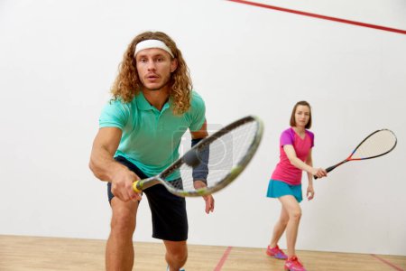 Téléchargez les photos : Jeune couple sportif pratiquant le squash sur le terrain du club sportif. Focus sur l'homme attrayant frapper balle avec raquette - en image libre de droit