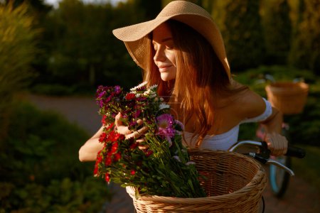 Téléchargez les photos : Charmante dame sentant les fleurs sauvages fleurit pendant la promenade à vélo dans le parc le week-end. Féminin, tendresse et charme de la jeunesse - en image libre de droit