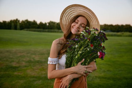 Téléchargez les photos : Belle femme tenant un bouquet de fleurs sauvages debout sur un champ de campagne verdoyant. Concept de féminité, beauté naturelle et innocence - en image libre de droit