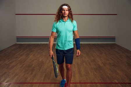 Photo for Portrait of man holding squash racquet walking among sport court. Confident racquetball player and professional sport - Royalty Free Image