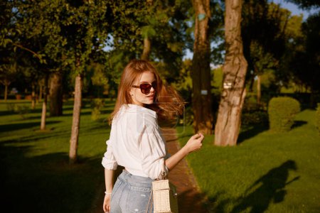 Téléchargez les photos : Portrait extérieur de jeune jolie femme marchant dans le parc à la journée ensoleillée. Heureuse adolescente joyeuse avec look fashion tournant avec son dos à la caméra - en image libre de droit