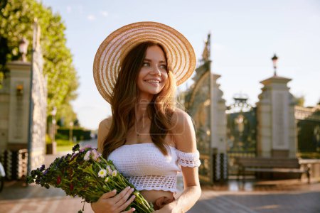 Photo for Portrait of gentle beautiful young woman holding wildflower bouquet in summer park. Charming romantic girl in fashion straw hat - Royalty Free Image