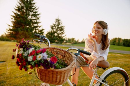 Téléchargez les photos : Jeune fleuriste hipster femme portant des écouteurs écoutant de la musique se reposer après une longue course à vélo pour les fleurs sauvages sur le terrain vert. Artisanat passe-temps et voyager à l'extérieur - en image libre de droit