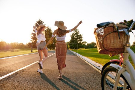 Photo for Young female friends enjoying road trip on bicycle spending weekend together. View on excited joyful teenager girls running on road and bike parked at sidewalk - Royalty Free Image