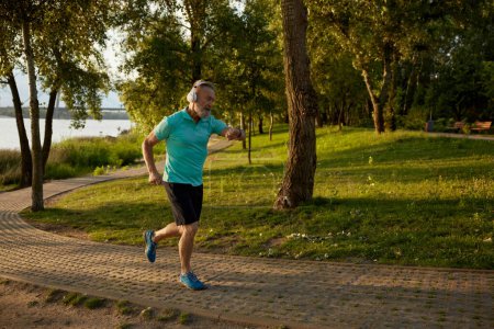 Photo for Senior man running in park during early morning. Elderly sportsman wearing headset looking at wristwatch fitness tracker caring for his health and wellbeing - Royalty Free Image