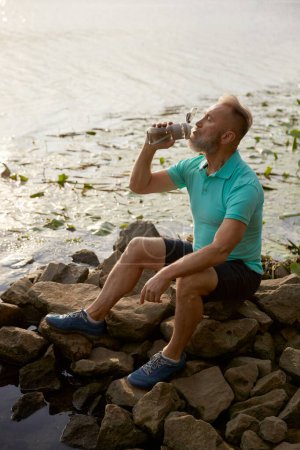 Photo for Old mature sportsman enjoying beautiful wild nature after morning jog. Senior man sitting on stone by pond with water lily flower drinking water taking rest - Royalty Free Image