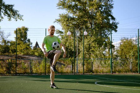 Téléchargez les photos : Joueur de football masculin frappant le ballon avec son entraînement de genou sur le terrain de football de rue. Loisirs sportifs en plein air pour adultes - en image libre de droit