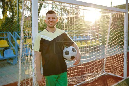 Photo for Relaxed football player with ball standing leaned at soccer gate on field outdoors. Tired sportsman feeling happy and satisfied looking at camera - Royalty Free Image