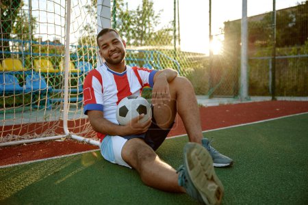 Photo for Relaxed football player with ball sitting leaned at soccer gate on field outdoors. Tired sportsman feeling happy and satisfied looking at camera - Royalty Free Image