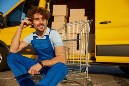 Photo for Smiling professional male delivery courier resting after handing to customer while sitting outdoors nearby cargo van - Royalty Free Image