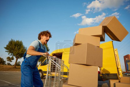 Photo for Stressed clumsy courier screaming looking at falling parcel boxes from fully loaded trolley cart. Accident at work - Royalty Free Image