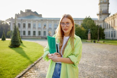 Photo for Outdoors portrait of smart female student wearing eyeglasses and casual clothes holding studying accessories standing over university campus yard on background - Royalty Free Image