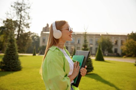 Foto de Retrato de vista lateral de una estudiante satisfecha que usa auriculares en el campus universitario sobre fondo borroso. Bastante lindo adolescente escuchando música o interesante podcast, estudiando el lenguaje - Imagen libre de derechos