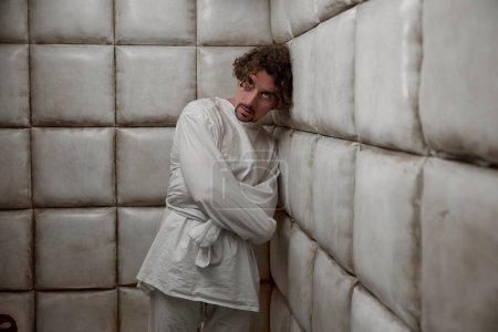 Photo for Calm man with schizophrenia mental disorder in white padded room. Male patient with madness on face wearing straitjacket standing leaning to soft walls in psychiatric ward - Royalty Free Image