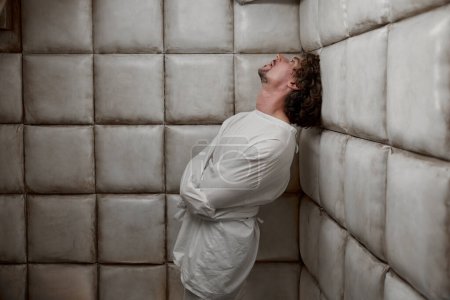 Photo for Calm man with schizophrenia mental disorder in white padded room. Male patient with madness on face wearing straitjacket standing leaning to soft walls in psychiatric ward - Royalty Free Image