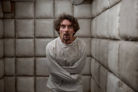 Foto de Hombre asustado con esquizofrenia trastorno mental en la habitación acolchada blanca. Paciente masculino con locura en la cara usando camisa de fuerza de pie en habitación blanda en la sala psiquiátrica - Imagen libre de derechos