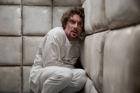 Foto de Loco hombre asustado paciente usando camisa de fuerza asustado sentado en la esquina de la habitación acolchada - Imagen libre de derechos