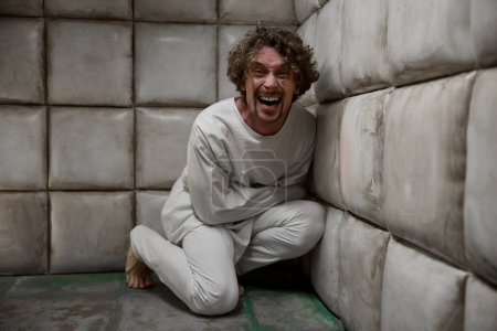Foto de Loco loco paciente con camisa de fuerza riendo aterrador sentado en la esquina de la habitación acolchada - Imagen libre de derechos