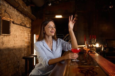 Photo for Positive young woman calling bartender while rest in bar after hard work day. Attractive businesswoman celebrating successful end of week drinking cocktail in restaurant or nightclub - Royalty Free Image