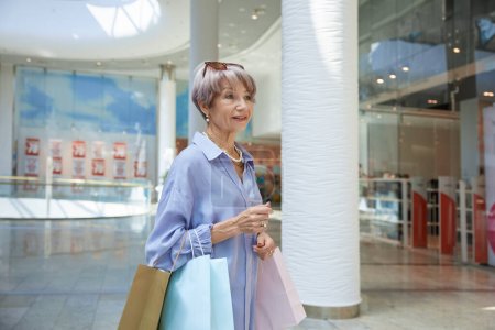 Téléchargez les photos : Portrait d'une séduisante femme âgée avec des sacs à provisions sur vitrine de magasin. Élégant grand-mère se sentant satisfait profiter de réductions dans la boutique - en image libre de droit