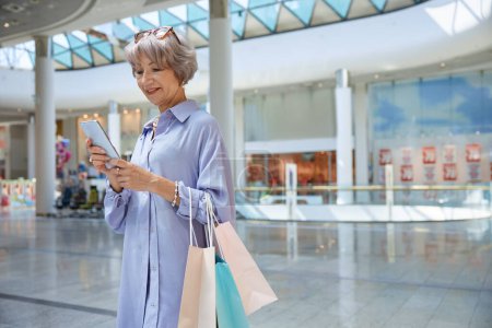 Foto de Atractiva mujer mayor con teléfono y bolsas de compras sobre escaparate de la tienda del centro comercial. Elegante abuela sintiéndose satisfecho disfrutando de descuentos en tienda - Imagen libre de derechos