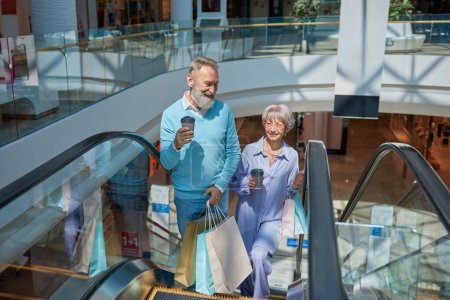 Photo for Senior couple with purchases in bags riding down shopping mall escalator enjoying the day - Royalty Free Image