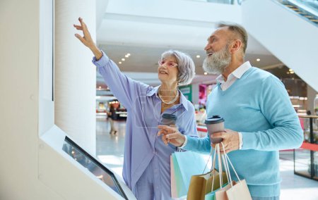 Photo for Aged smiley married couple standing at self-service kiosk in shopping mall. Casual senior man and woman enjoying sale discount using modern technology for purchases - Royalty Free Image
