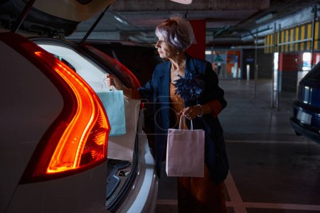 Téléchargez les photos : Heureuse dame âgée satisfaite de charger les achats dans des sacs à provisions dans le coffre de la voiture au parking - en image libre de droit