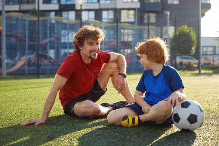 Foto de Padre joven enseñando a su hijo pequeño a jugar al fútbol diciendo reglas. Padres felices y niños sentados en el campo de fútbol al aire libre - Imagen libre de derechos