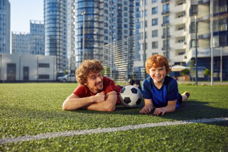Téléchargez les photos : Portrait d'un père enjoué et d'un petit garçon joyeux allongé sur de l'herbe de terrain de football. Happy loisirs sportifs en famille le week-end - en image libre de droit