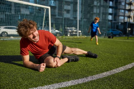 Photo for Young father soccer player injured his leg after falling during training match with son. Sportive injury and trauma on field - Royalty Free Image
